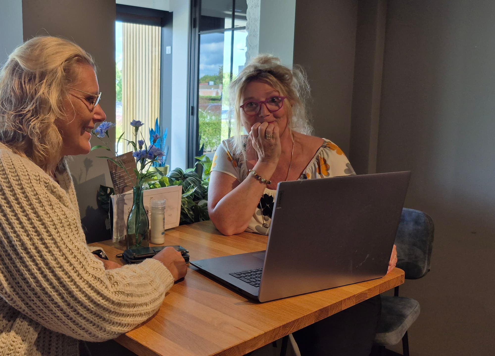 Two women sitting at a table with a laptop, engaged in a relaxed coaching conversation in a bright, cozy space.