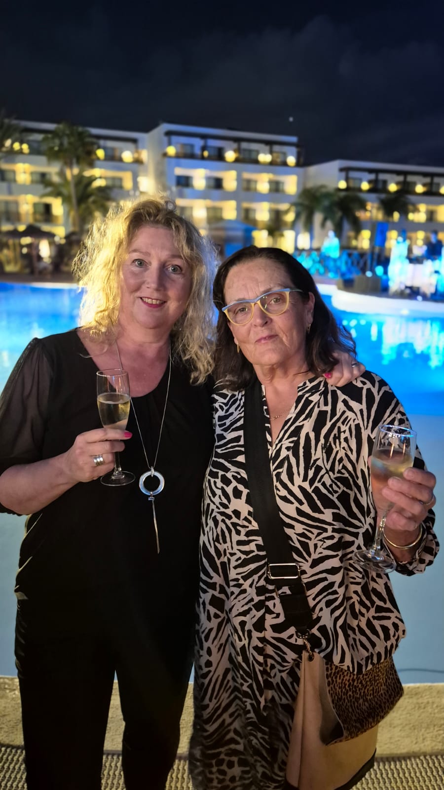 Two women standing close together at an evening event by a lit-up pool, smiling and holding glasses of sparkling wine